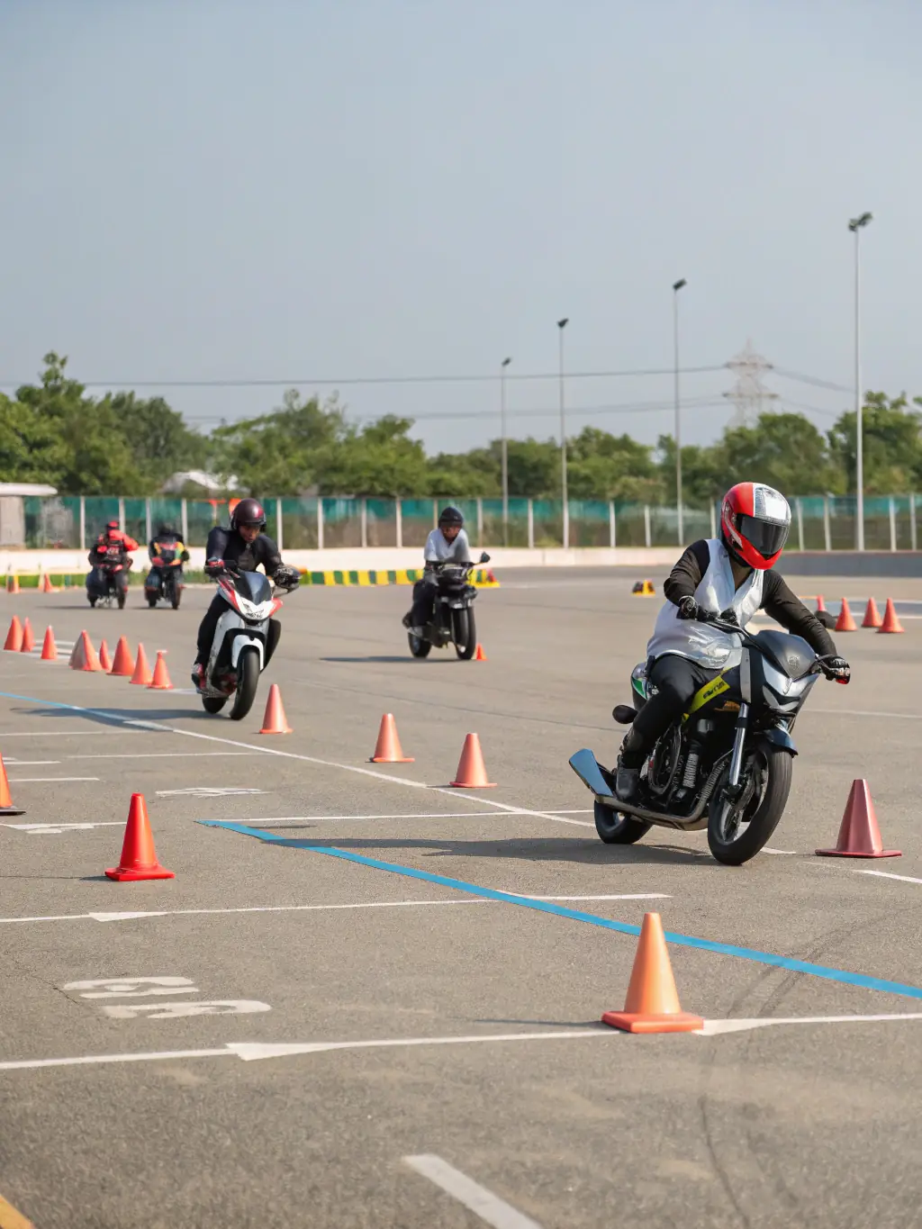 A motorcycle rider in full gear practicing maneuvers on a training course, cones and instructor visible in the background, sunny day.